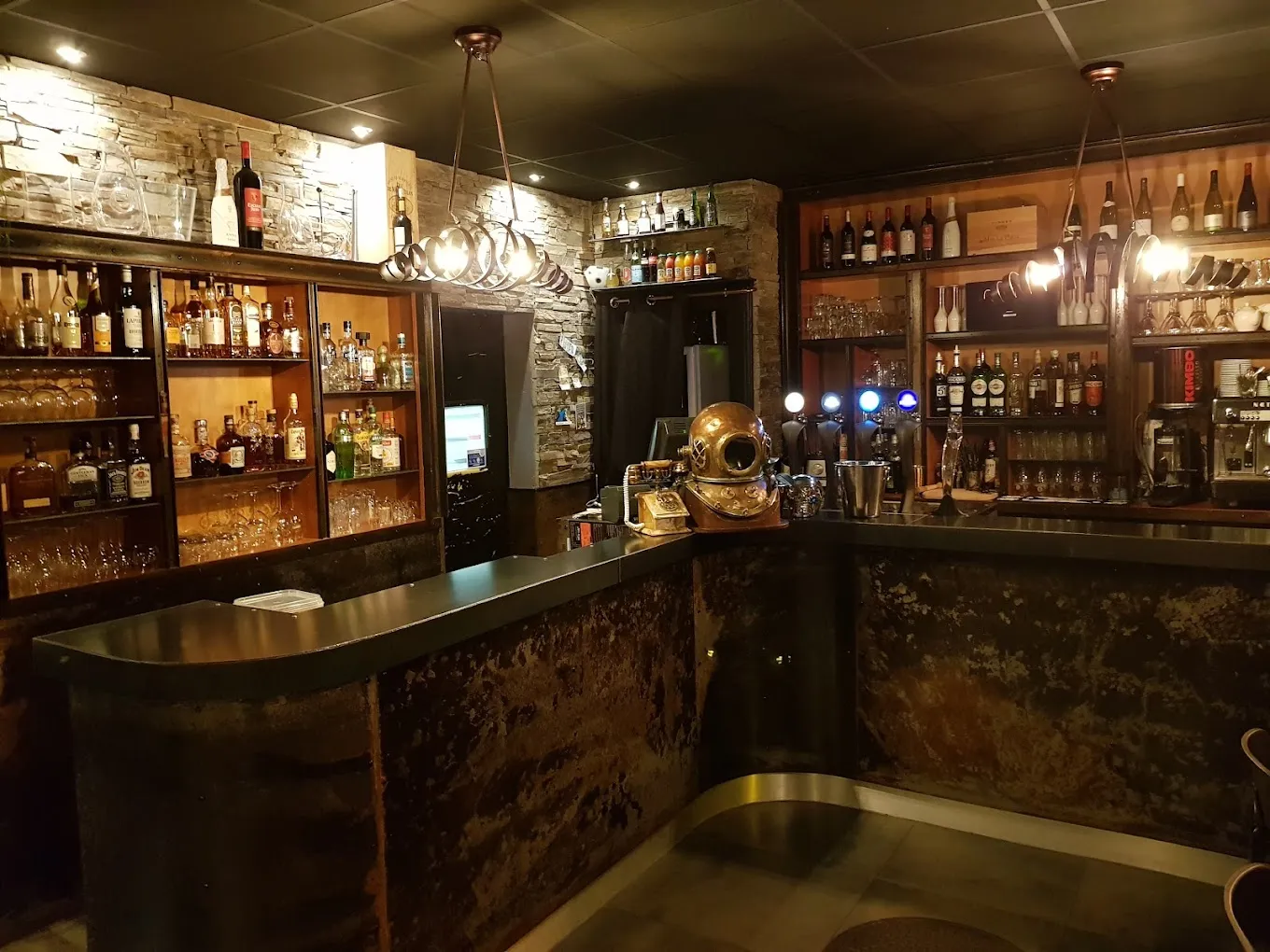 Le bar du restaurant en soirée, mur en pierre et étagères de bouteilles.
